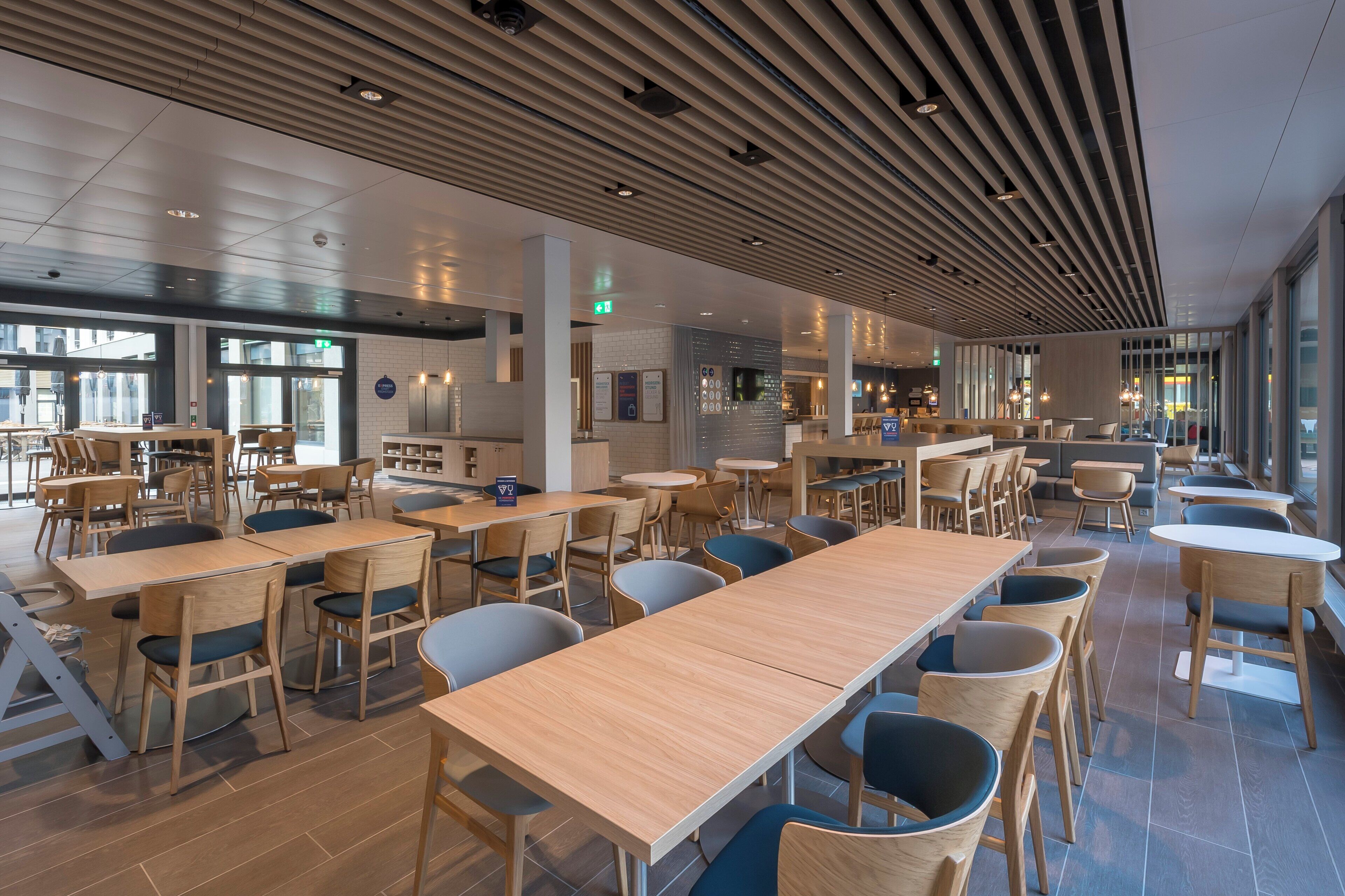 Un vaste espace dédié à la salle à manger, avec de nombreuses tables et chaises en bois, de grandes fenêtres et un design moderne au niveau du plafond.