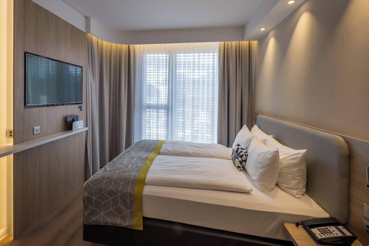 Een hotelkamer met een bed, een aan de muur bevestigde televisie, een raam met gordijnen en een nachtkastje met telefoon.