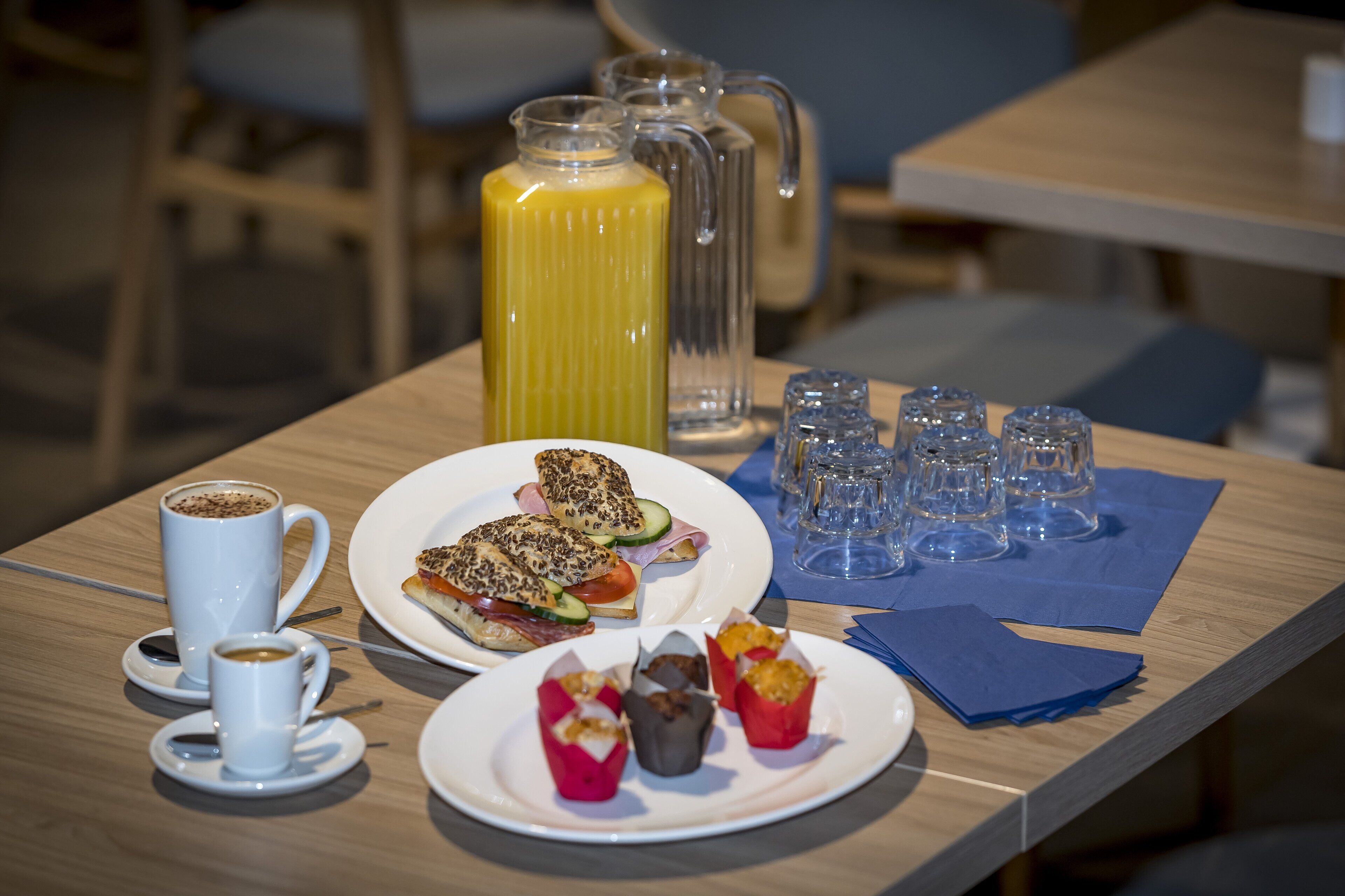 Une table de petit-déjeuner avec une tasse de café, un pichet de jus d’orange, une assiette de sandwichs et une assiette de viennoiseries.