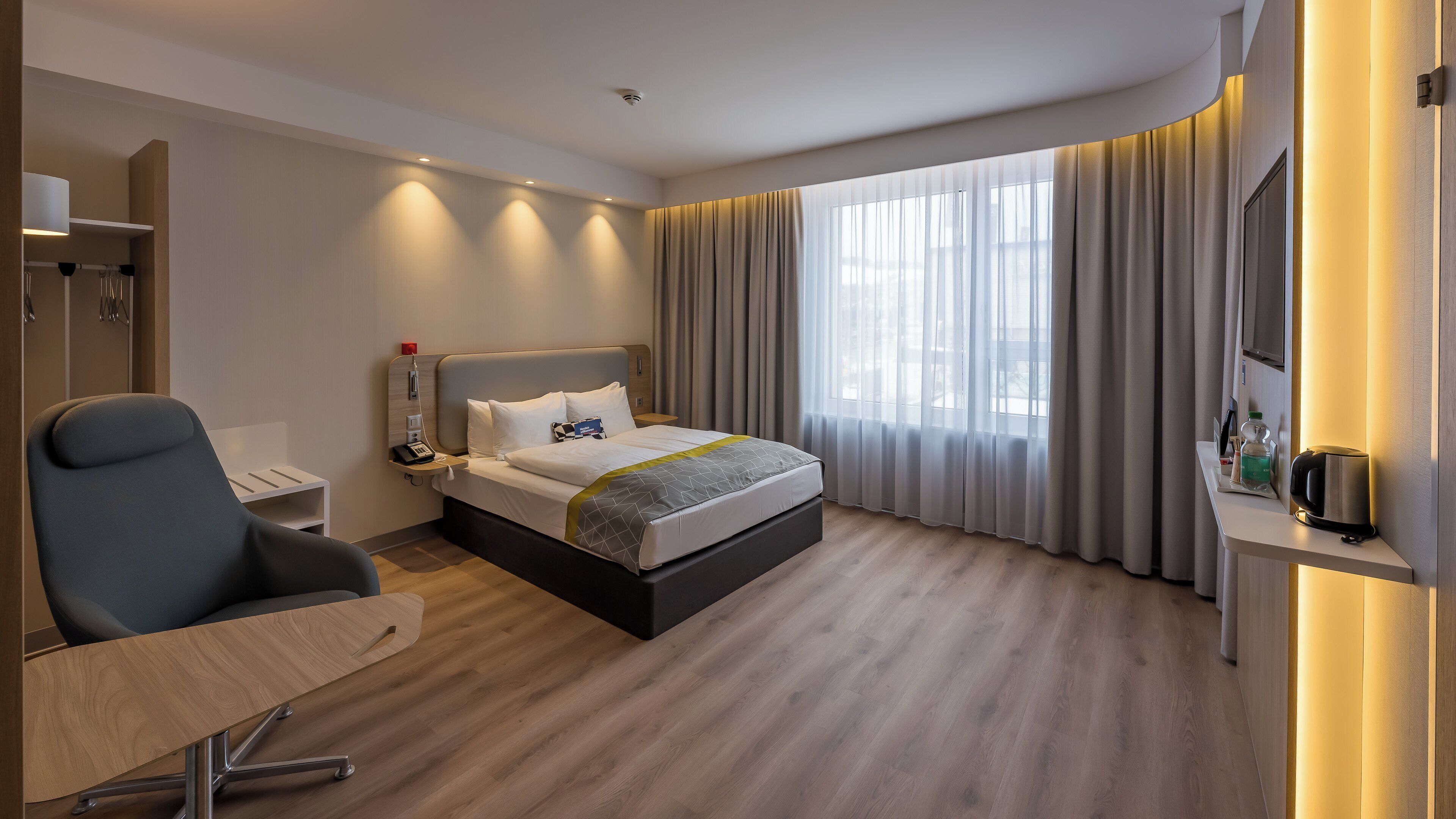 Et moderne hotelværelse med seng, skrivebord, stol, natbord og fjernsyn.