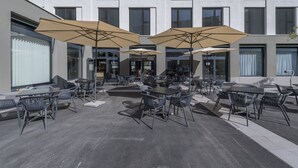 Un espace extérieur aménagé avec des tables et des chaises, à l’ombre de grands parasols, devant un bâtiment moderne doté de nombreuses fenêtres.