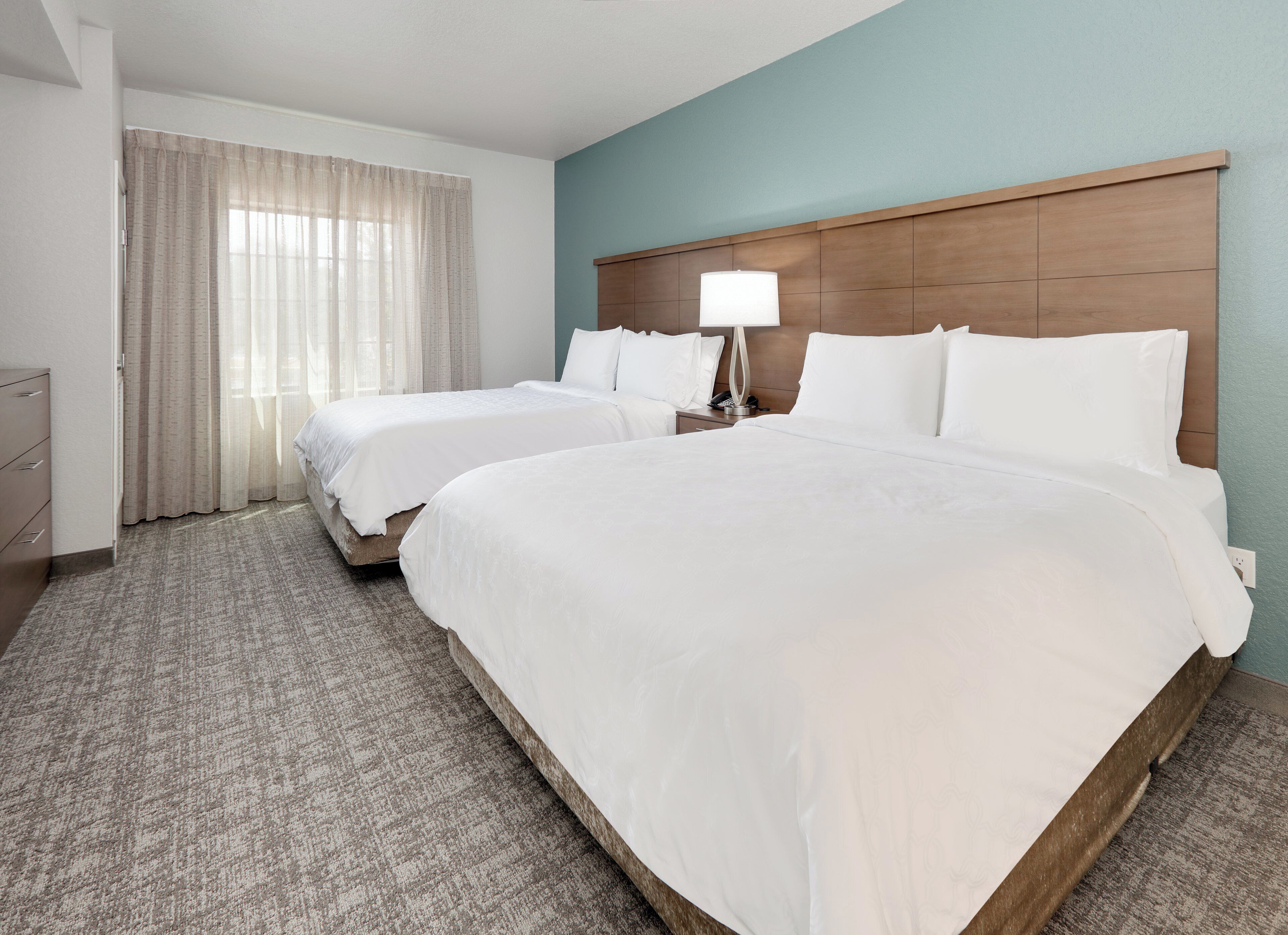 Une chambre d’hôtel avec deux lits, une table de chevet avec une lampe, une commode et une fenêtre avec des rideaux.