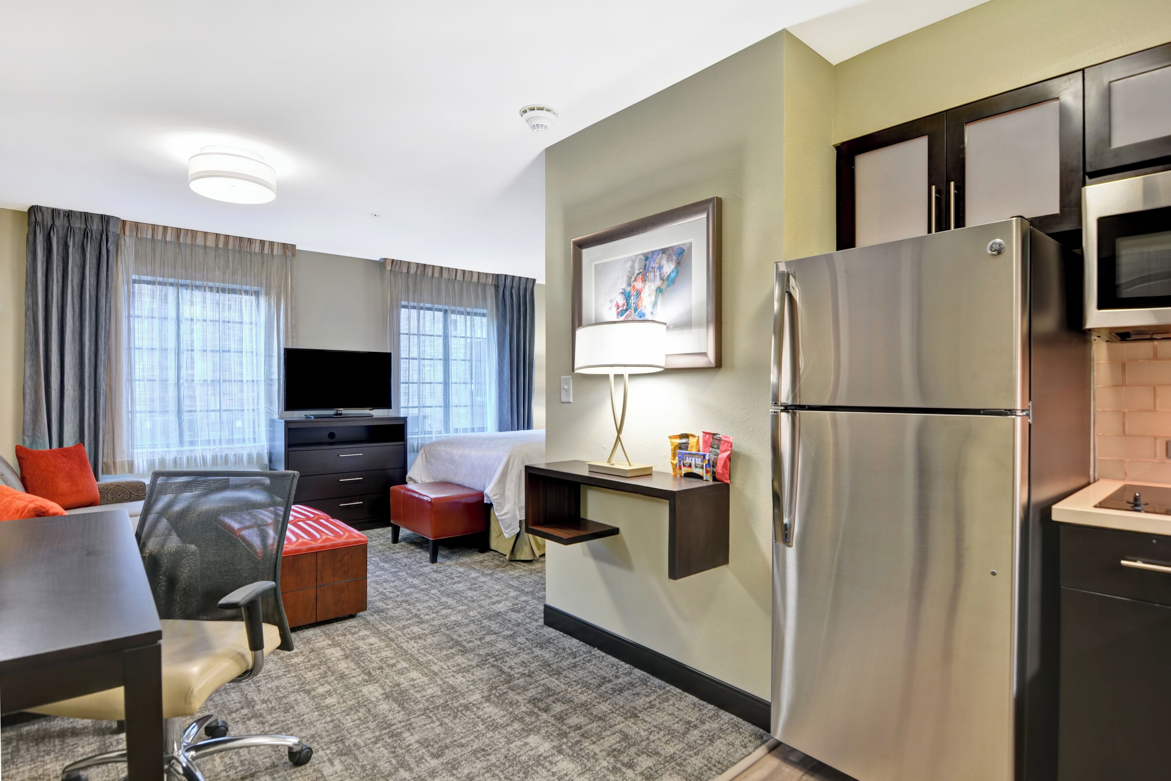Quarto de hotel com cozinha compacta, cama, televisão e escrivaninha com cadeira.