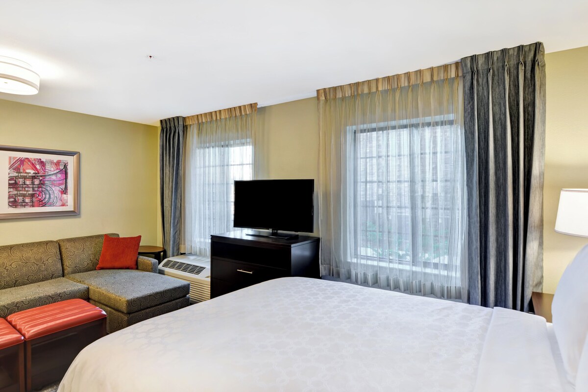 Quarto de hotel com cama, sofá, televisão e janela com cortinas.