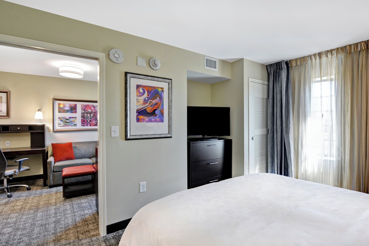 Quarto de hotel com cama, escrivaninha, cadeira, televisão, obras de arte e cortinas.
