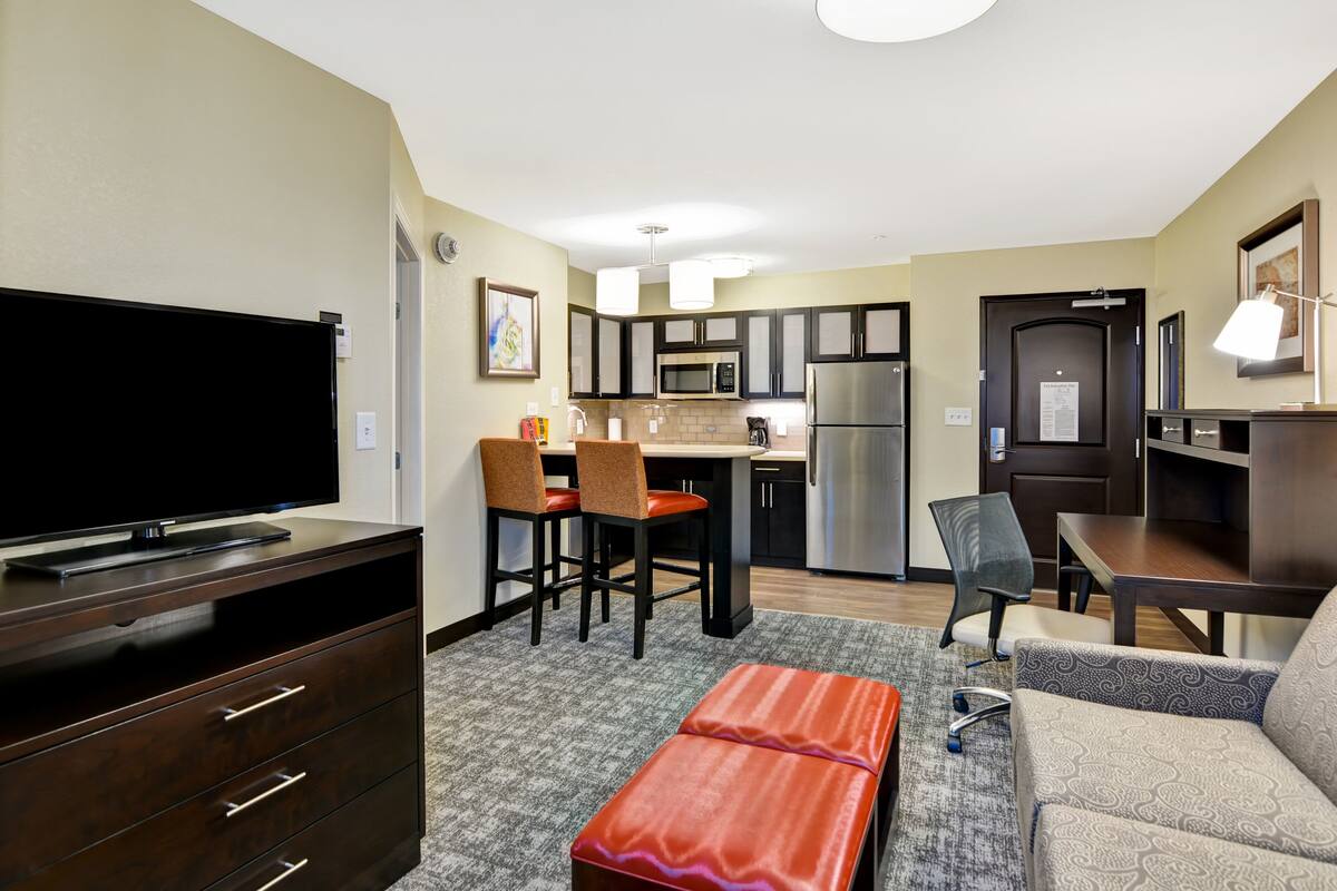 A compact hotel room with a kitchenette, a flat-screen TV, a sofa, a dining table, and a small red bench.