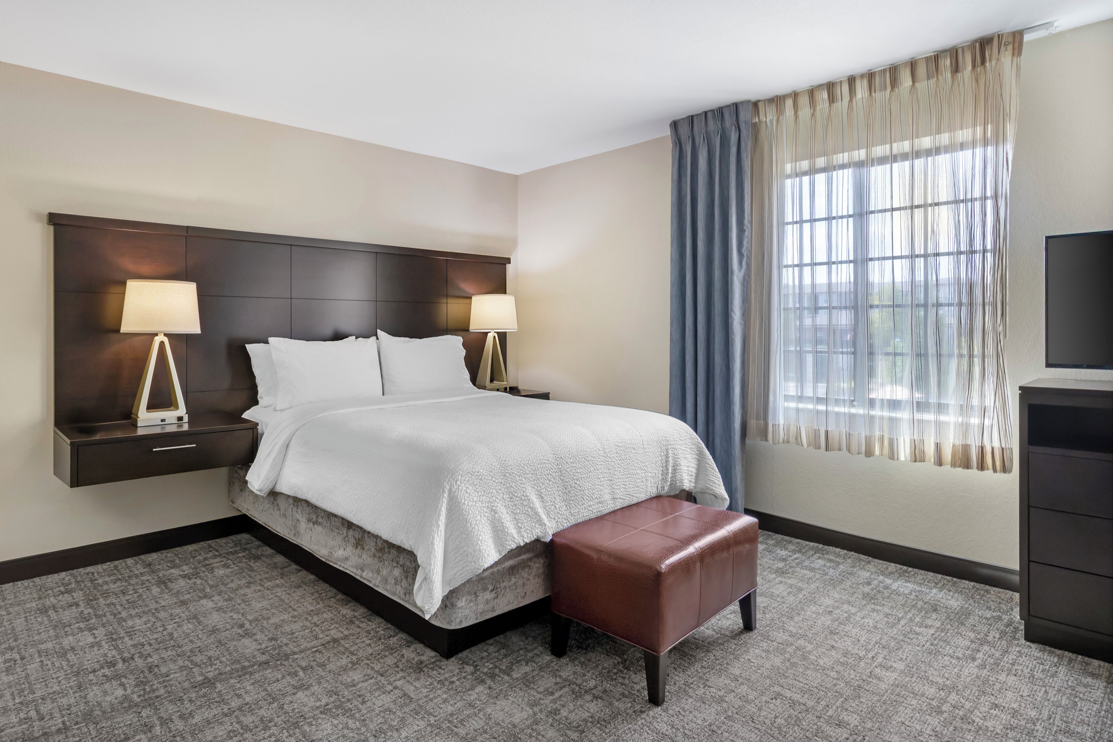 A hotel room with a bed, bedside tables, lamps, a TV, a window with curtains, and a carpeted floor.