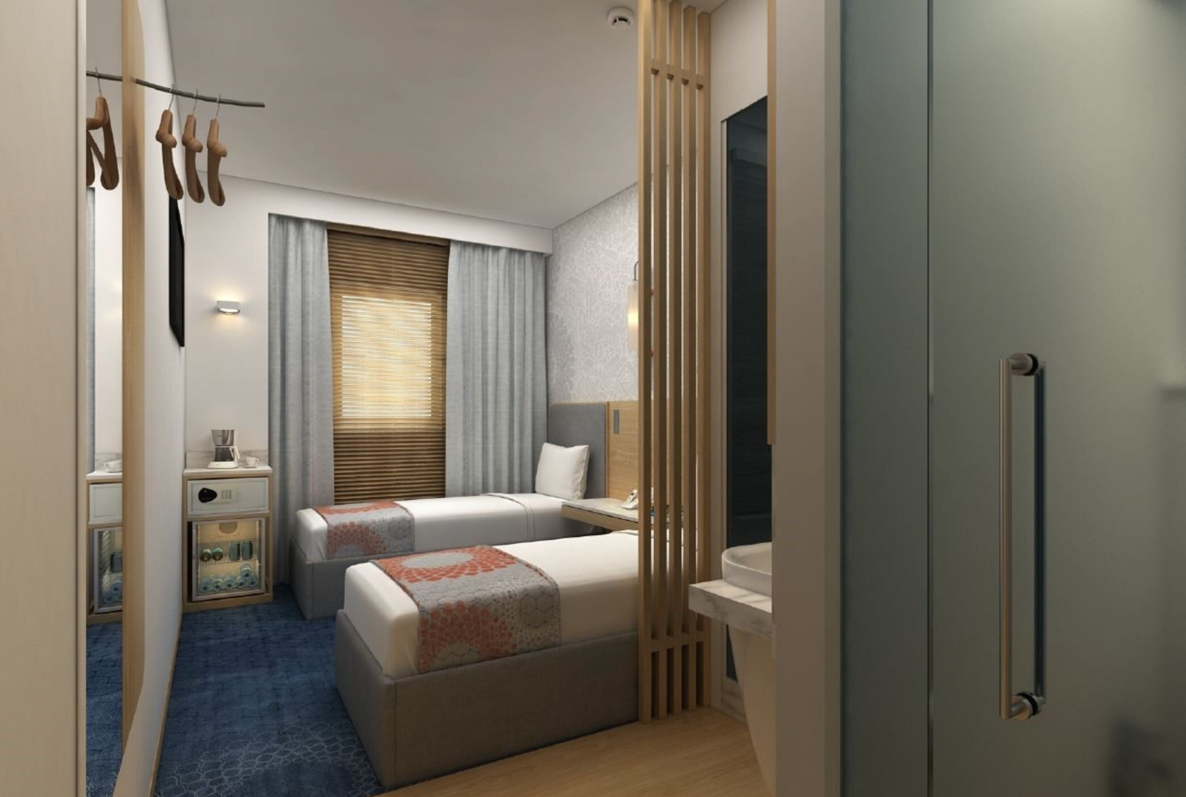 A hotel room with two beds, a small bedside table, a nightstand, a window with blinds, a wall-mounted lamp, and a bathroom with a sink and mirror.