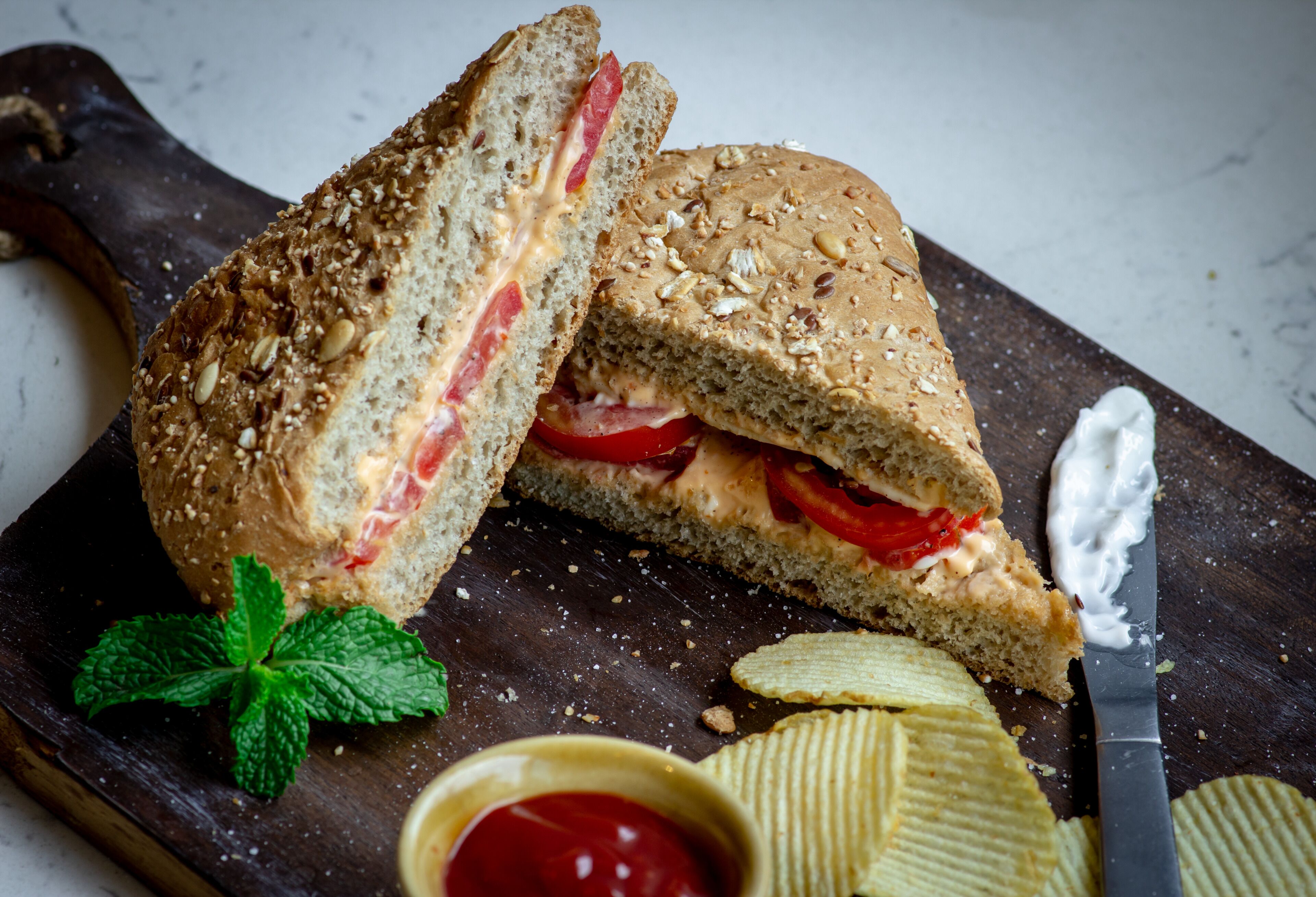 Un sándwich cortado por la mitad sobre una tabla de madera, con un cuchillo y un tazón pequeño de kétchup.
