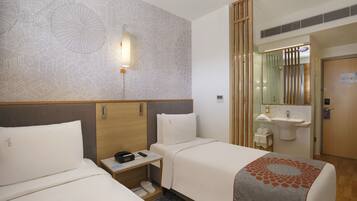 A hotel room with two beds, a bedside table with a phone, a small table with a lamp, and a bathroom with a sink and mirror.