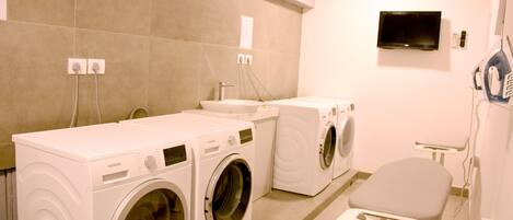 A laundry room with three white washing machines, a sink, and a chair.