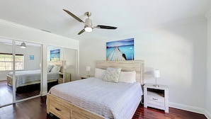A bedroom with a bed, a nightstand, a mirror, a ceiling fan, and a painting on the wall.