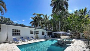 A swimming pool with lounge chairs and umbrellas, surrounded by palm trees and a building with windows.