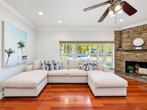 A living room with a sectional sofa, a ceiling fan, a fireplace, and a large window with a view of palm trees.