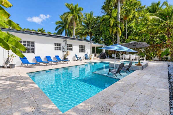 A swimming pool with lounge chairs and umbrellas, surrounded by palm trees and a white building.