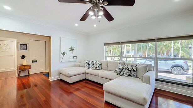 A spacious living room with a large sectional sofa, a ceiling fan, and a view of the street through large windows.