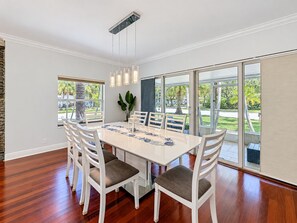 A dining area with a large table, white chairs, and a view of a garden through large windows.