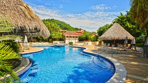 A swimming pool with a thatched roof structure, lounge chairs, and a tropical garden setting. - Los Suenos Resort Colina 8E (Jacó)