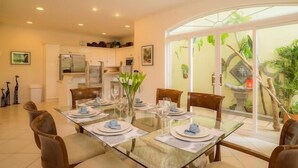 A dining area with a glass table, chairs, and a view of a garden through a glass door. - Los Suenos Resort Colina 8E (Jacó)