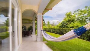 A covered patio with a hammock, a glass door leading to a dining area, and a well-manicured lawn. - Los Suenos Resort Colina 8E (Jacó)