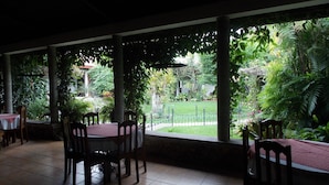 Un espacio cubierto para comer al aire libre con mesas y sillas, rodeado de abundante vegetación y vistas a un jardín.