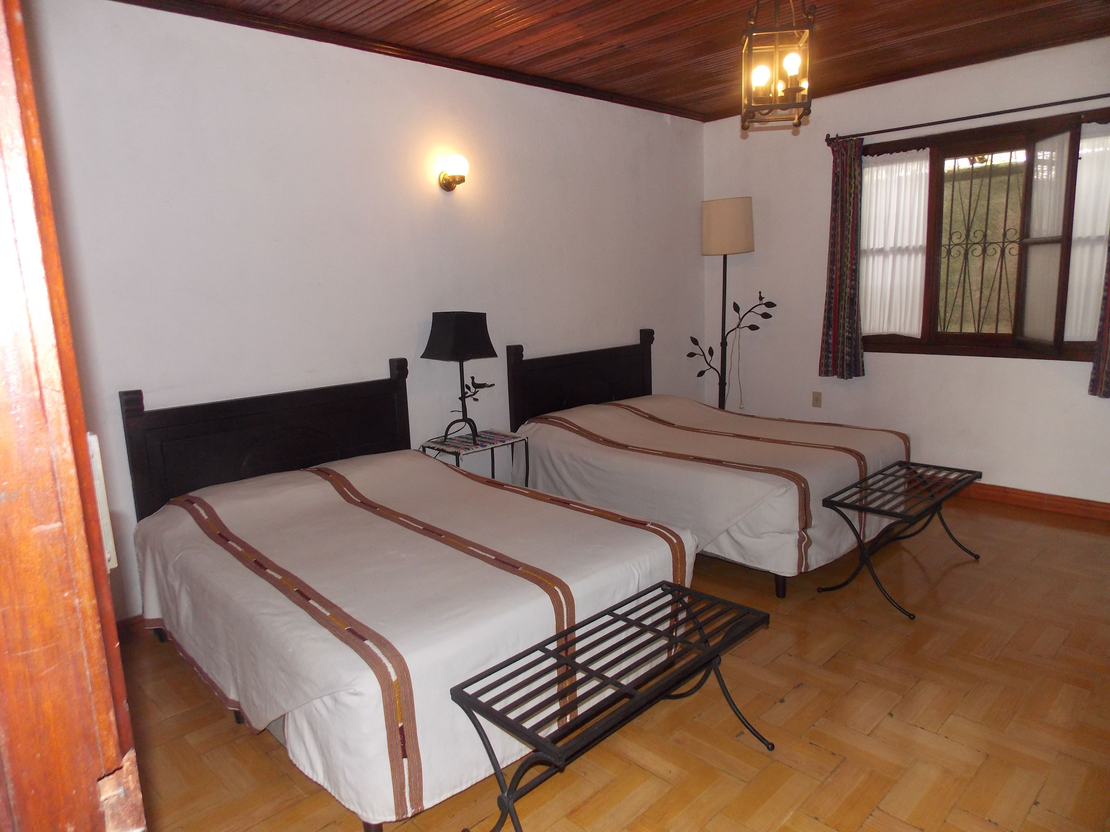 A room with two beds, a small table, a lamp, a wall-mounted light, and a window with curtains.