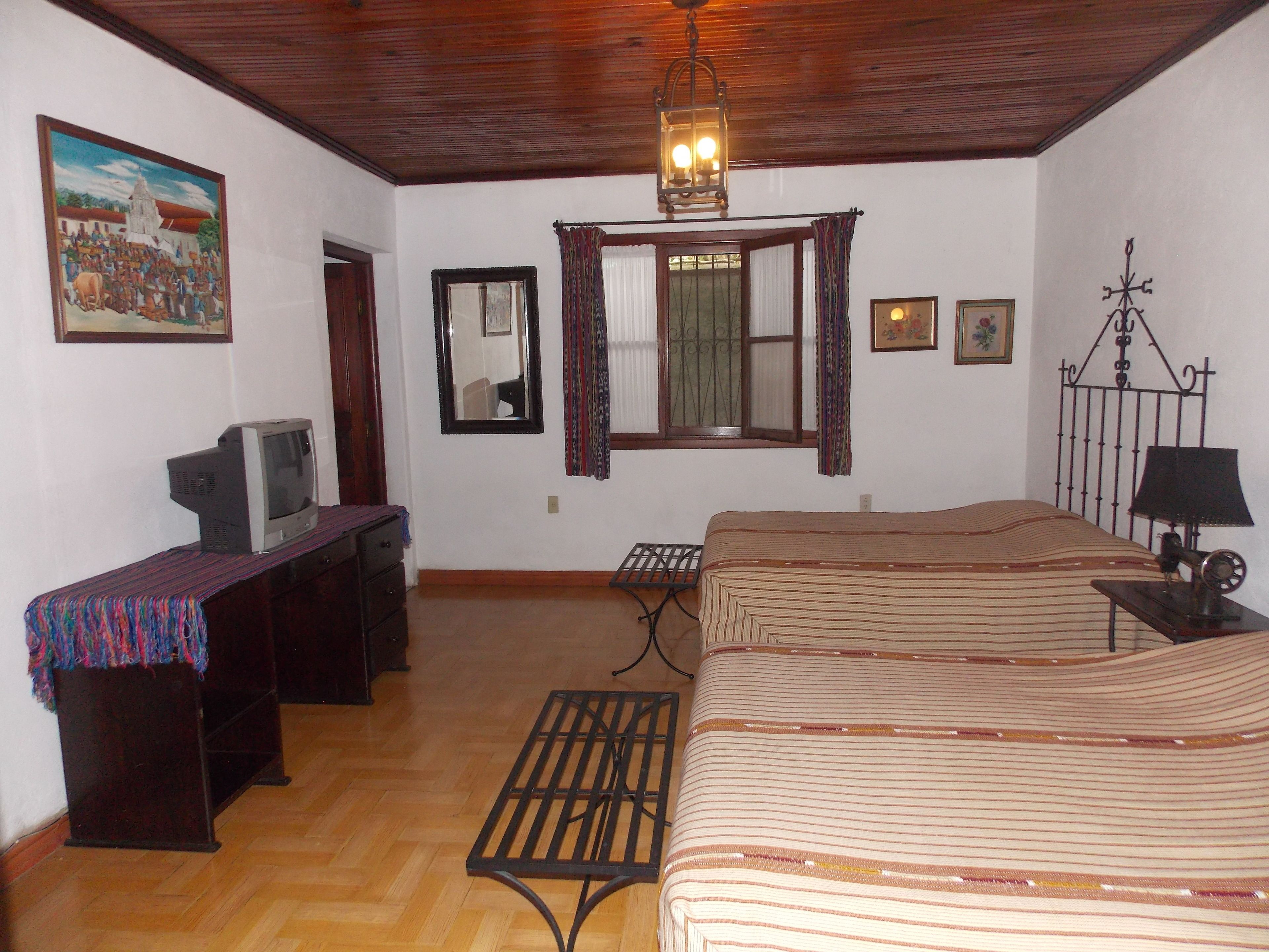 A bedroom with a bed, a desk, a television, a mirror, and a hanging light fixture.