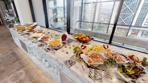 A buffet table with various food items and utensils, set in a room with large windows overlooking a cityscape. - Estelar Square (Medellín)
