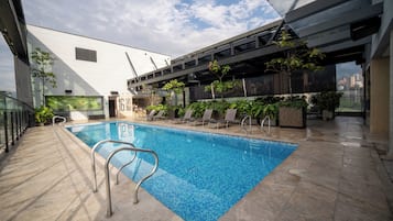 Una piscina interior con vistas al horizonte urbano, una zona techada con sillones de descanso y suelo de baldosas.