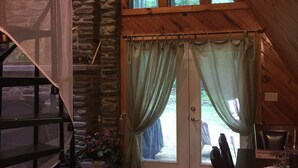 L’intérieur chaleureux d’un chalet avec une cheminée en pierre, des poutres en bois, un coin repas et une grande fenêtre donnant sur la verdure.