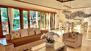 Un salon spacieux avec un grand canapé, une table basse et deux fauteuils. De grandes fenêtres offrent une vue sur un jardin luxuriant.