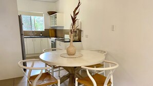 Een ronde eettafel met witte stoelen en een houten onderstel, omgeven door een witte muur en een raam.