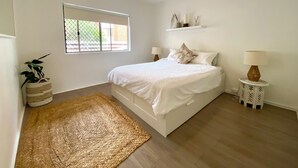 Een slaapkamer met een bed, een raam, een plafondventilator, een nachtkastje met een lamp en een geweven vloerkleed.