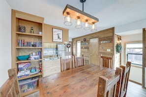 Ein Essbereich mit Holztisch und Stühlen, ein Bücherregal mit diversen Gegenständen und eine Küche im Hintergrund.