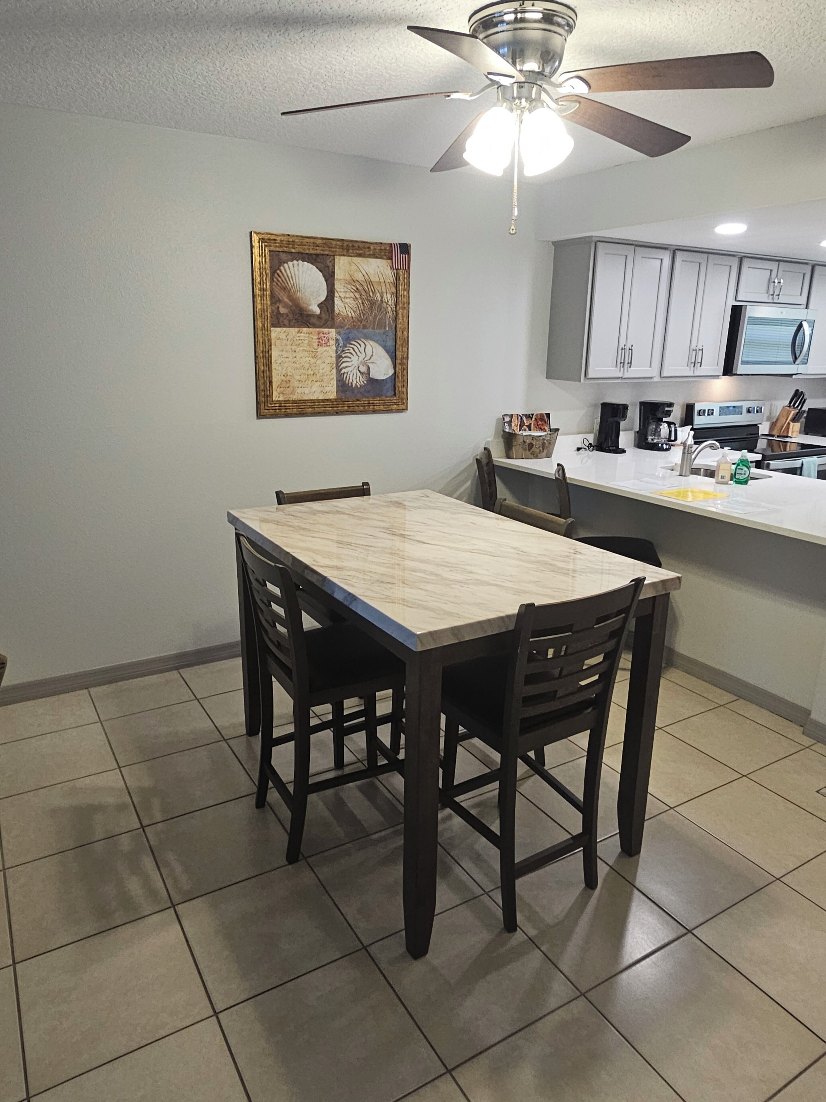 A dining area with a wooden table and chairs, a ceiling fan, and a kitchen in the background.