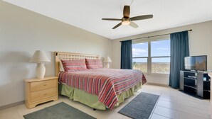A bedroom with a bed, two bedside tables, a ceiling fan, a TV, and a large window with curtains. - Oceanfront Condo, Family Friendly Close To Orlando Attractions (Cocoa Beach)