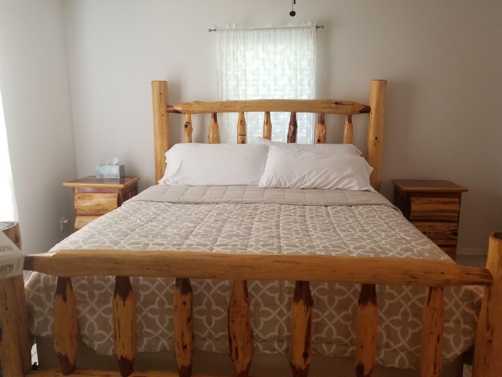 A wooden bed with a patterned bedspread, two bedside tables, and a window with sheer curtains.
