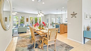 A spacious dining area with a wooden table and chairs, a chandelier, and a view of the outdoors through large windows. - The Sun Dial | Waterfront Home on Siesta Key w/ Private Pool & Large Backyard, Close to Beach! (Siesta Key)
