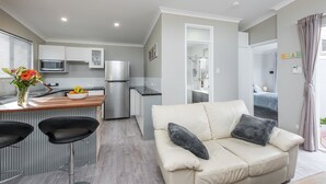 A modern living room with a beige sofa, two black chairs, a wooden dining table, and a kitchen area with a refrigerator, microwave, and a bowl of fruit. - Prestigious Northshore Guest House - Comfortable Living Close To Mullaloo Beach (Kallaroo)