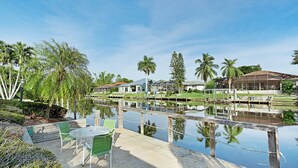 A serene outdoor area with a concrete patio, green chairs, and a table. A wooden dock extends over a calm water body, surrounded by lush greenery and palm trees. - Villa Tara: Gulf-Access, Heated Pool, Private Dock (Cape Coral)