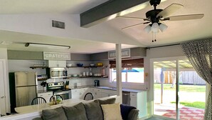 Une cuisine moderne avec des armoires blanches, un réfrigérateur et un four à micro-ondes. On y trouve un coin repas avec une table en verre et un canapé. La pièce est équipée d’un ventilateur de plafond et offre une vue sur le jardin arrière grâce à une porte-fenêtre coulissante.
