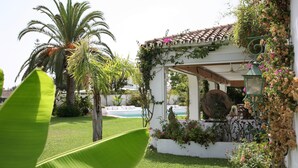 A covered outdoor seating area with a pool, surrounded by lush greenery and flowering plants.