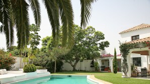 A swimming pool surrounded by a well-maintained lawn, palm trees, and a white building with a tiled roof.