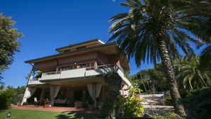 A two-story house with a balcony, surrounded by lush greenery and palm trees.