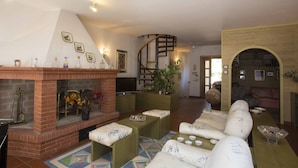 A spacious living room with a brick fireplace, comfortable seating, a television, and a spiral staircase.