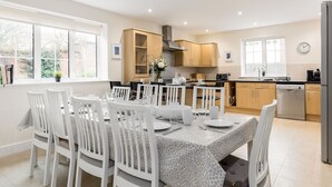 A modern kitchen with white cabinetry, a dining area with white chairs and a patterned tablecloth, and a large window providing natural light.