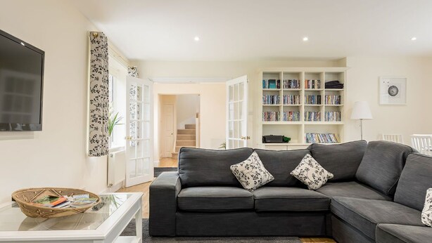 A modern living room with a dark grey sectional sofa, a glass coffee table, and a white side table. A bookshelf filled with books is visible in the background.