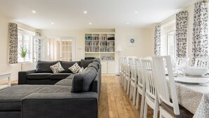 A modern living room with a grey sofa, a dining area with white chairs and a table, and a bookshelf filled with books.