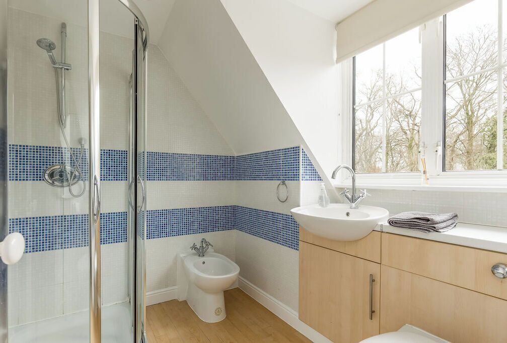 A bathroom with a glass shower enclosure, a bidet, a sink, and a window with a view of trees.