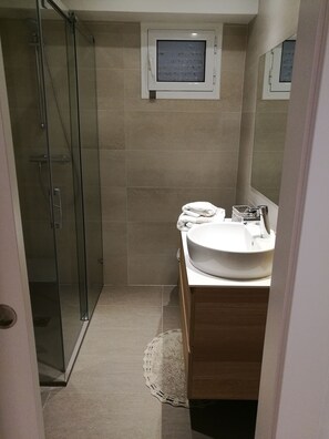 Un baño moderno con cabina de ducha de vidrio, un lavamanos blanco y una pequeña ventana sobre el espejo.